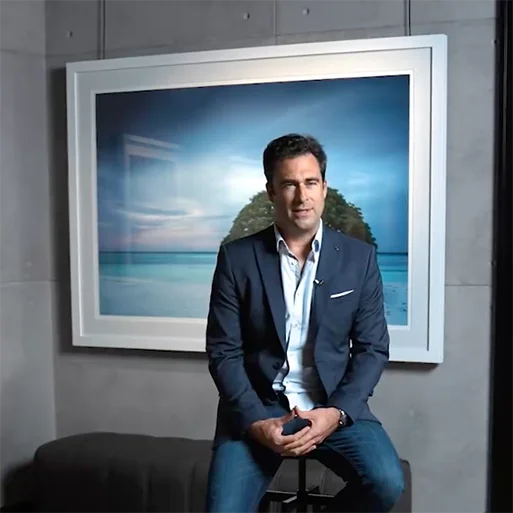 Photographer Anthony Lamb seated on a stool in front of an exhibition print at Gallery X